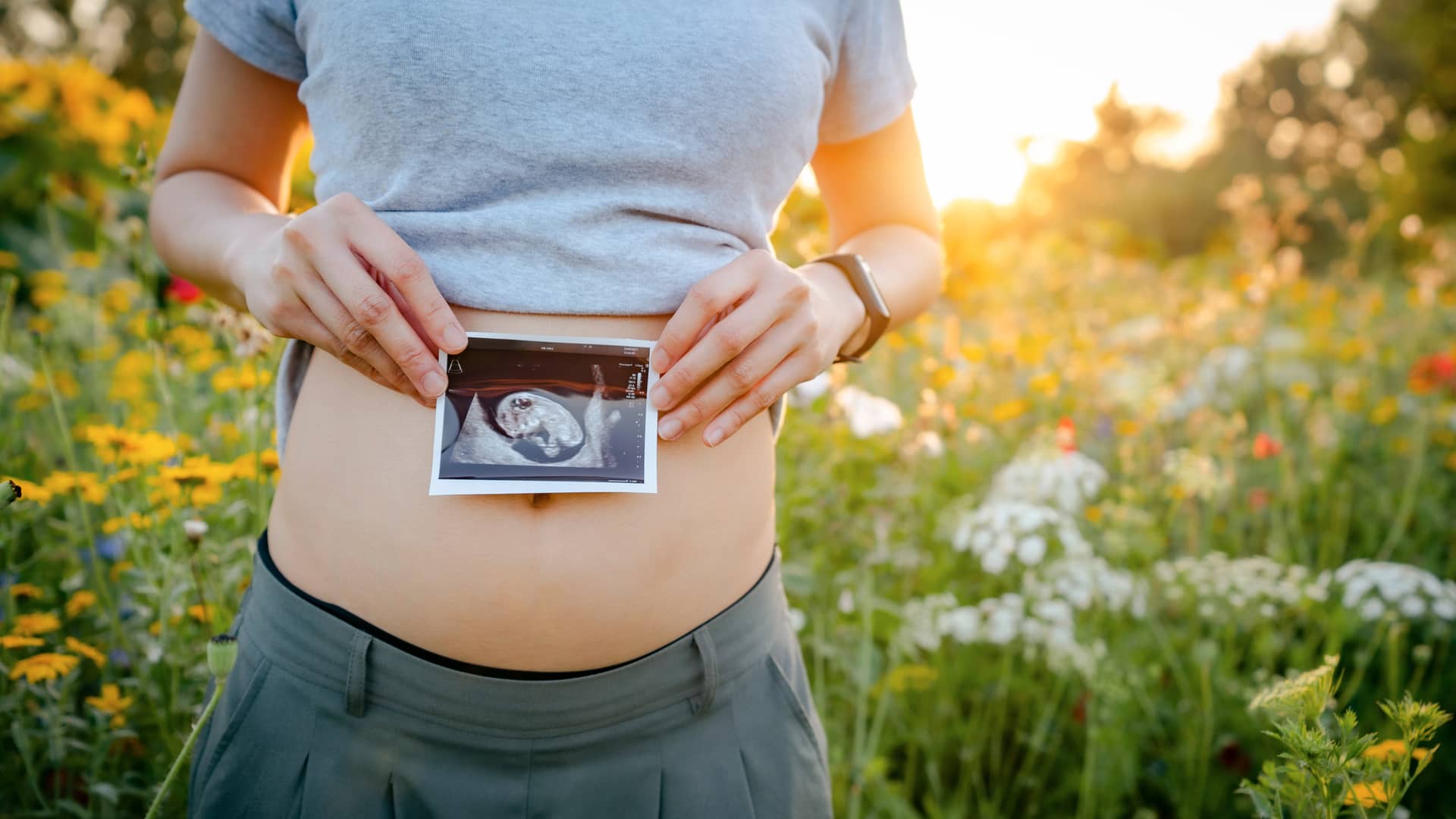 Una joven en el campo luciendo su pequeña barriga de embarazada de la semana 13 junto a una ecografía de su bebé