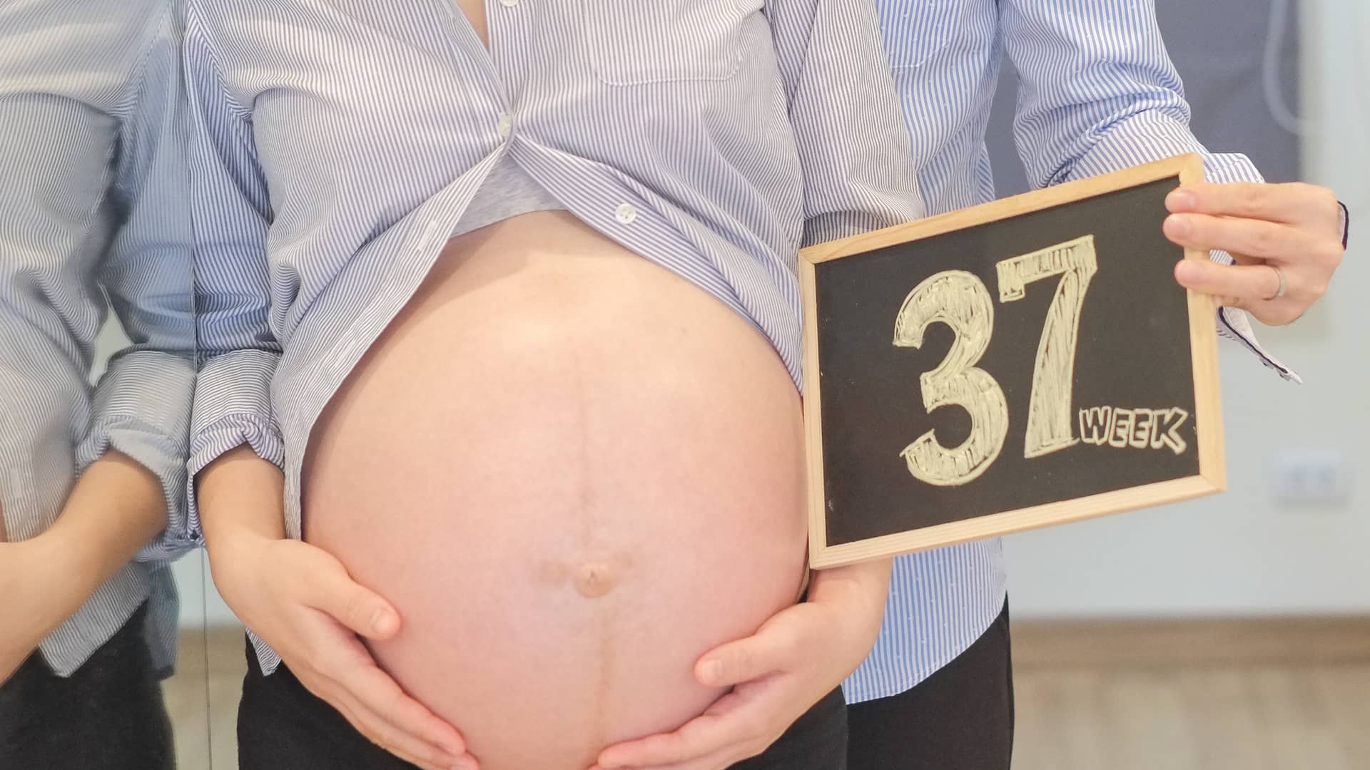 Mujer con la barriga de embarazada descubierta y mostrando una pequeña pizarra que muestra el número 37 haciendo referencia a su semana de embarazo